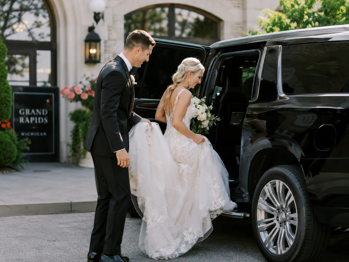 wedding couple stepping into a black Chevy Suburban or Mercedes-Benz Sprinter van, dressed in wedding attire, outside a Grand Rapids venue